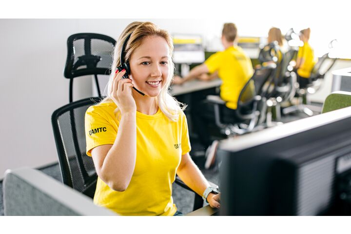 Auf dem Bild sieht man eine Frau in einem kurzärmeligen gelben T-Shirt. Am Ärmels steht ÖAMTC. Sie sitzt vor einem Bildschirm und hat ein Headset auf.