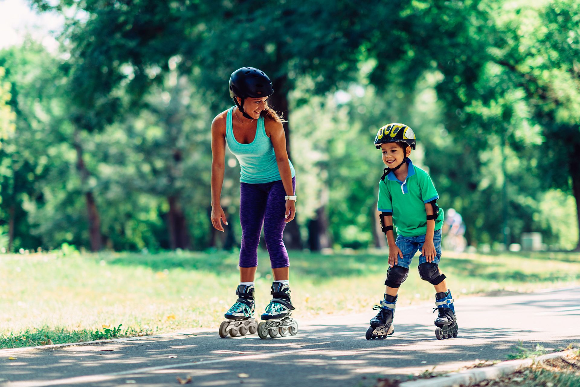 Mutter und Sohn beim Rollschuhlaufen bzw. Inlineskaten im Park. Beide tragen Helm, der Junge auch Knie- und Ellbogenschützer.