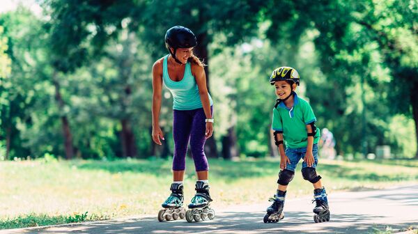 Mutter und Sohn beim Rollschuhlaufen bzw. Inlineskaten im Park. Beide tragen Helm, der Junge auch Knie- und Ellbogenschützer.
