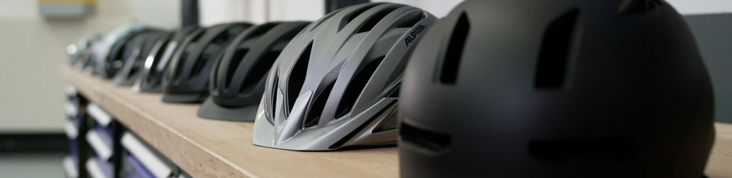 Mehrere Fahrradhelme in verschiedenen Farben und Designs sind in einer Reihe auf einer Werkbank angeordnet. 