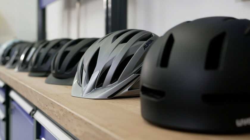 Mehrere Fahrradhelme in verschiedenen Farben und Designs sind in einer Reihe auf einer Werkbank angeordnet. 