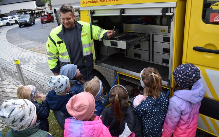 Pannenfahrer Thomas steht vor einer Gruppe an Kindergartenkinder und deutet auf den Inhalt seines Pannenfahrzeuges. 