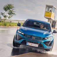 Ein älterer Herr am Steuer eines blauen Autos weicht einer Wasserfontäne bei einem Fahrsicherheitskurs aus