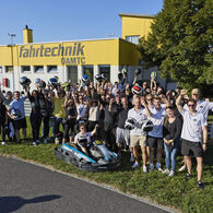 Eine Gruppe von Menschen steht versammelt hinter einem Go-Kart. Einige halten einen Helm in die Höhe. Im Hintergrund sieht man ein Gebäude der ÖAMTC Fahrtechnik.