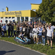 Eine Gruppe von Menschen steht versammelt hinter einem Go-Kart. Einige halten einen Helm in die Höhe. Im Hintergrund sieht man ein Gebäude der ÖAMTC Fahrtechnik.