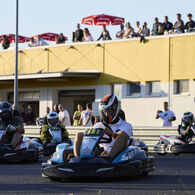 Vier Kart-Fahrer sind auf einer Rennstrecke zu sehen, im Hintergrund ein Gebäude mit Dachterasse, auf der Menschen dem Rennen zuschauen.