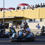 Vier Kart-Fahrer sind auf einer Rennstrecke zu sehen, im Hintergrund ein Gebäude mit Dachterasse, auf der Menschen dem Rennen zuschauen.