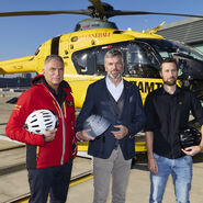 Drei Männer vor einem gelben Hubschrauber. Jeder der drei Männer hat einen Fahrradhelm in der Hand.
