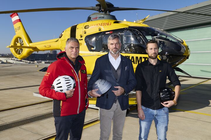 Drei Männer vor einem gelben Hubschrauber. Jeder der drei Männer hat einen Fahrradhelm in der Hand.