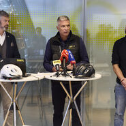 Drei Männer stehen vor einer Glaswand. Sie sprechen bei einer Pressekonferenz, auf einem Stehtisch vor Ihnen sind Mikrofone platziert.