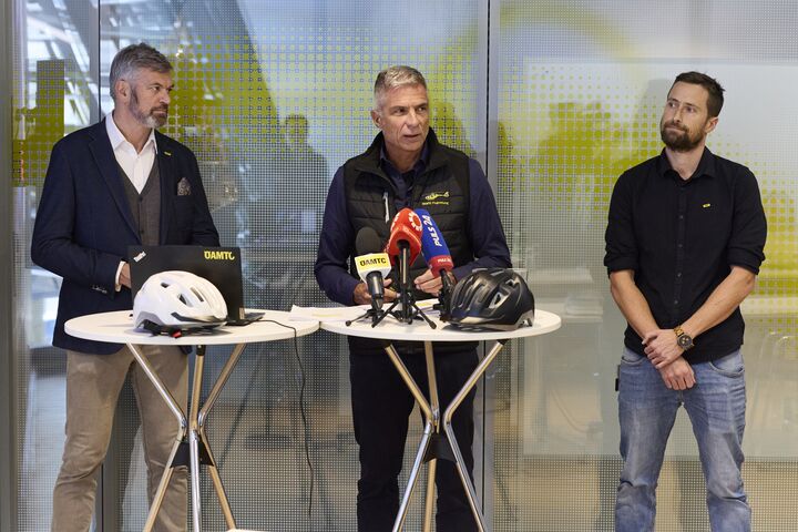 Drei Männer stehen vor einer Glaswand. Sie sprechen bei einer Pressekonferenz, auf einem Stehtisch vor Ihnen sind Mikrofone platziert.