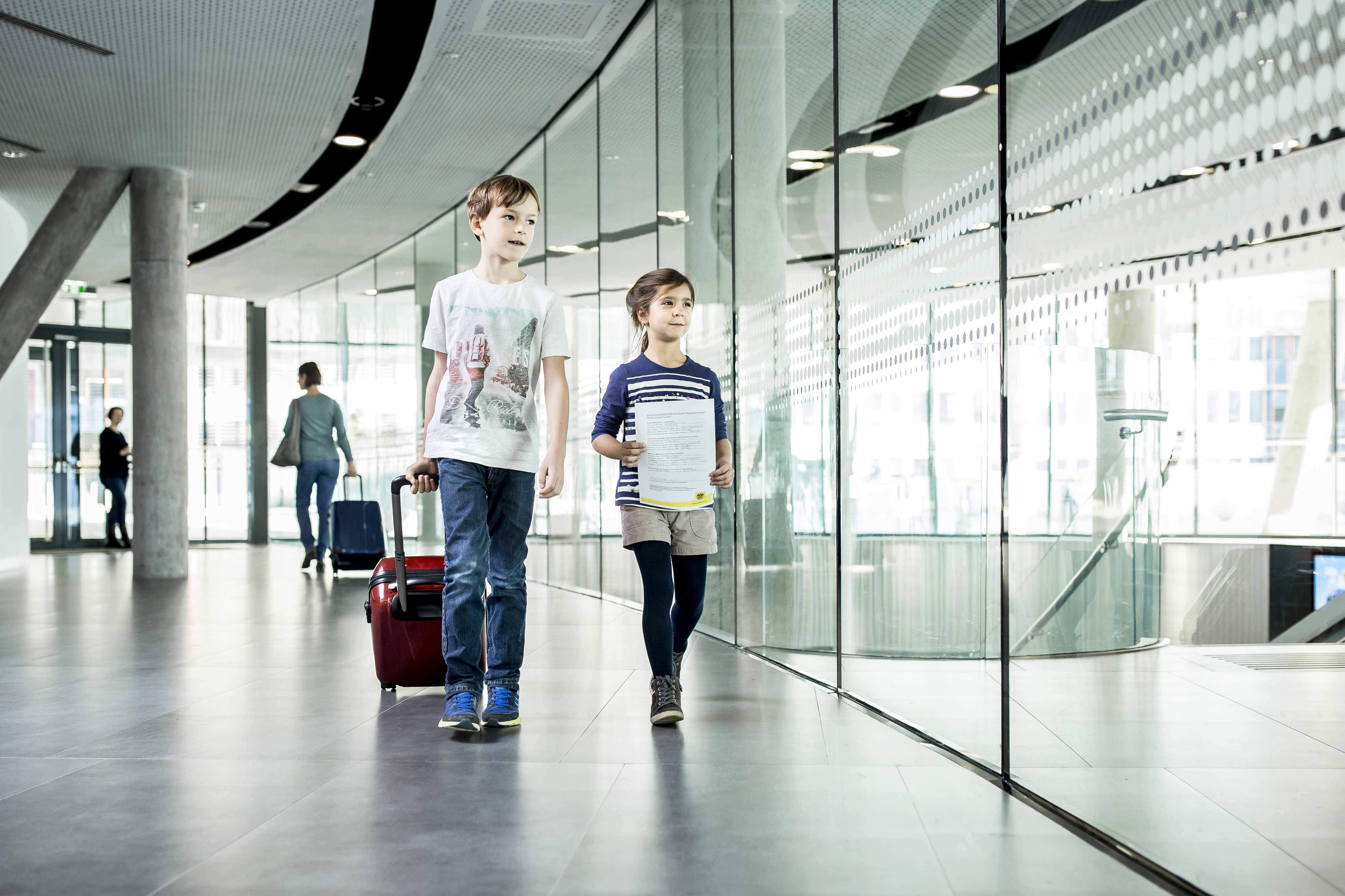 ein kleines Mädchen und ein kleiner Junge gehen einen Gang entlang. Der Junge zieht einen Trolley, das Mädchen hat ein Dokument in der Hand.