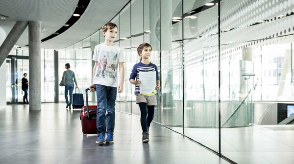 ein kleines Mädchen und ein kleiner Junge gehen einen Gang entlang. Der Junge zieht einen Trolley, das Mädchen hat ein Dokument in der Hand.