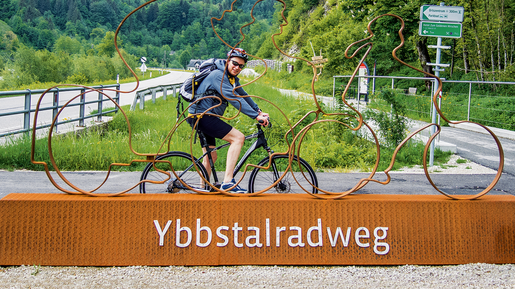 Das Bild zeigt den auto touring-Redakteur, der auf dem Ybbstal-Fahrradweg unterwegs ist und vor einem großen orangefarbenen Sockel posiert, auf dem "Ybbstalradweg" steht. Auf dem Sockel befindet sich auch eine Metallskulptur von zwei Radfahrern. Im Hintergrund sieht man eine Straße in grüner Landschaft mit Bäumen und Hügeln. 