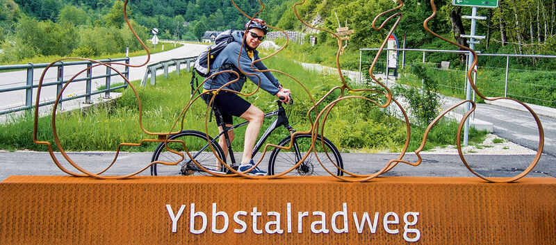 Das Bild zeigt den auto touring-Redakteur, der auf dem Ybbstal-Fahrradweg unterwegs ist und vor einem großen orangefarbenen Sockel posiert, auf dem "Ybbstalradweg" steht. Auf dem Sockel befindet sich auch eine Metallskulptur von zwei Radfahrern. Im Hintergrund sieht man eine Straße in grüner Landschaft mit Bäumen und Hügeln.