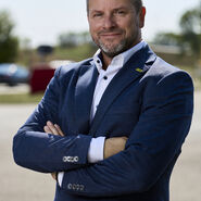 Porträt von Andreas Rouschal in einem blauen Anzug mit verschränkten Händen in die Kamera lächelnd, im Hintergrund sieht man ein Fahrtechnik-Training