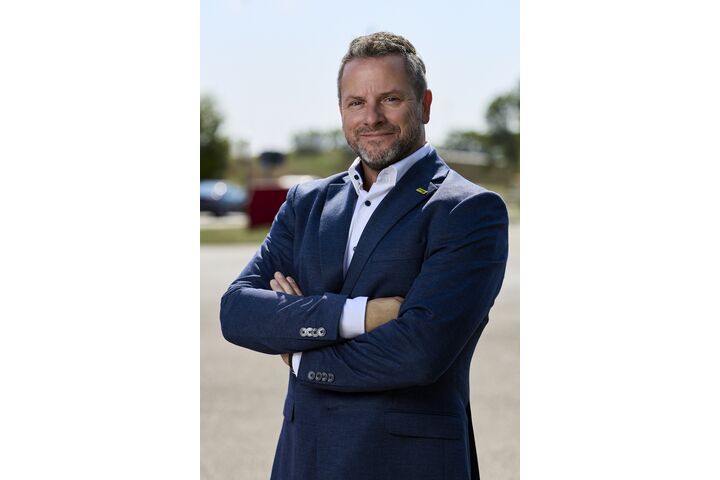 Porträt von Andreas Rouschal in einem blauen Anzug mit verschränkten Händen in die Kamera lächelnd, im Hintergrund sieht man ein Fahrtechnik-Training