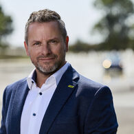 Porträt von Andreas Rouschal in einem blauen Anzug mit weißem Hemd, in die Kamera lächelnd, im Hintergrund sieht man ein Fahrtechnik-Training