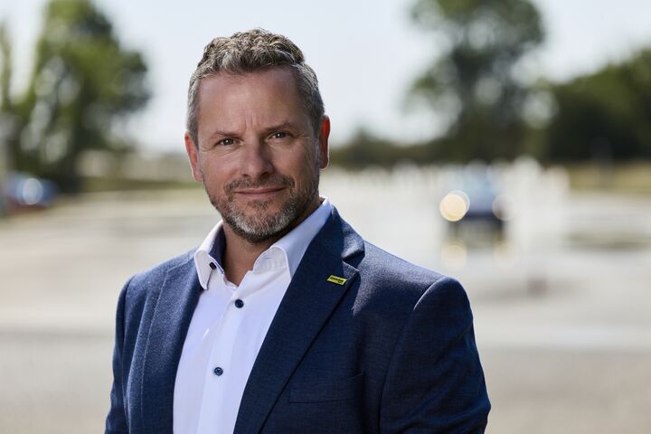Porträt von Andreas Rouschal in einem blauen Anzug mit weißem Hemd, in die Kamera lächelnd, im Hintergrund sieht man ein Fahrtechnik-Training