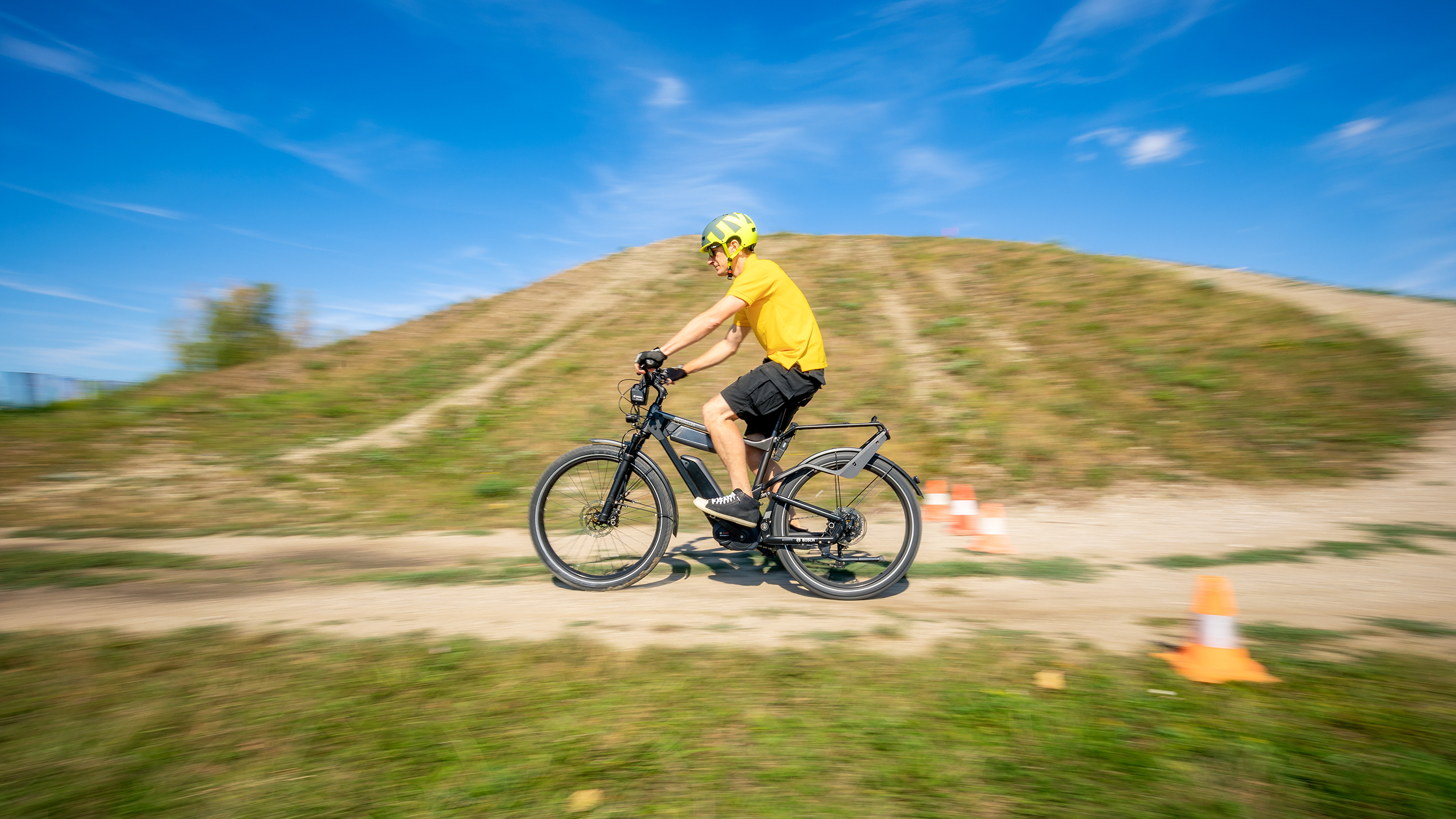 Der auto touring- Redakteur fährt mit einem E-Bike bei sommerlichem Wetter auf einem hügeligen Feldweg. An der Seite des Weges sind Pylonen aufgestellt. 