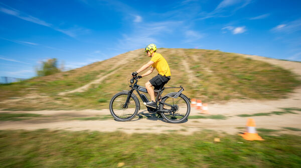 Der auto touring- Redakteur fährt mit einem E-Bike bei sommerlichem Wetter auf einem hügeligen Feldweg. An der Seite des Weges sind Pylonen aufgestellt.