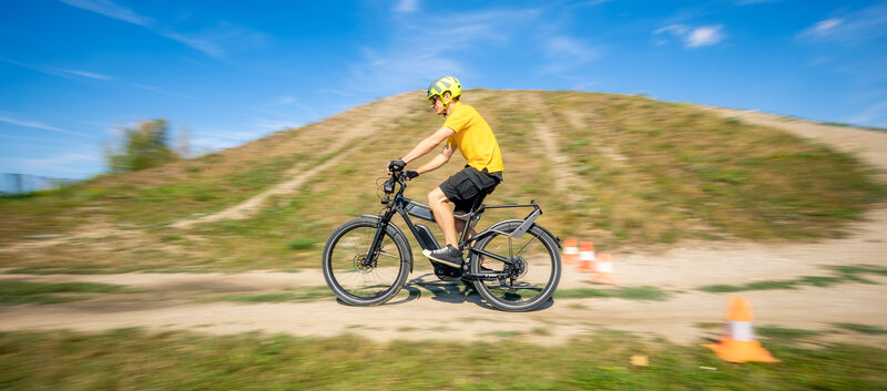 Der auto touring- Redakteur fährt mit einem E-Bike bei sommerlichem Wetter auf einem hügeligen Feldweg. An der Seite des Weges sind Pylonen aufgestellt.