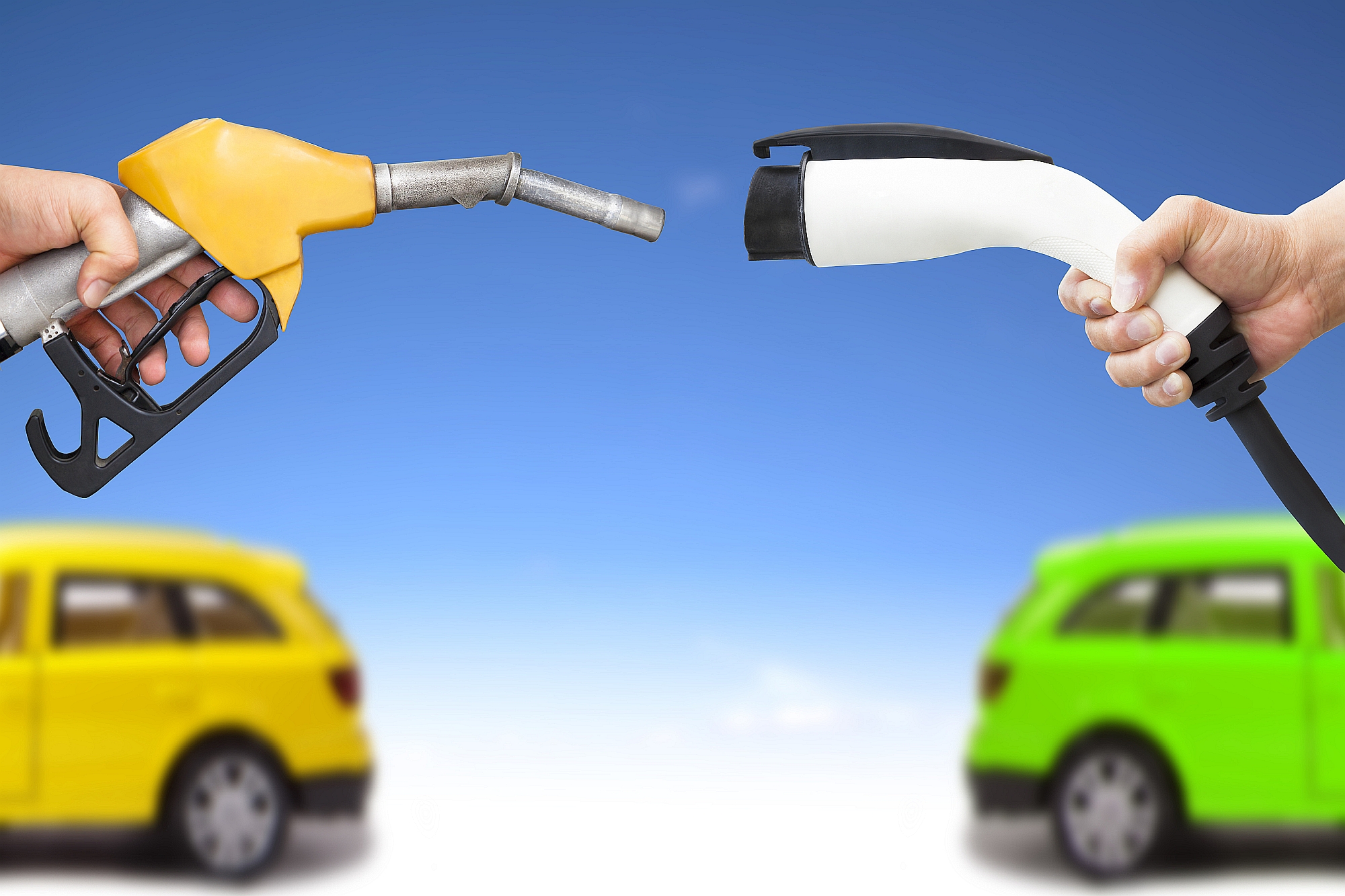 Ein gelber Treibstoff-Zapfhahn und ein weißes Ladekabel für ein Elektroauto zeigen zueinander, im Hintergrund sind ein gelbes und ein grünes Auto.