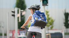 Ein Schulkind steht mit einem Fahrrad vor einer roten Ampel. Man sieht es von hinten. Es trägt einen Helm und einen blauen schulrucksack.