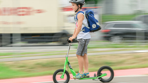 Ein Junge fährt auf einem grünen Tretroller, auch Scooter genannt, auf einem Weg neben einer Straße. Er trägt einen Helm und eine blaue Schultasche am Rücken.