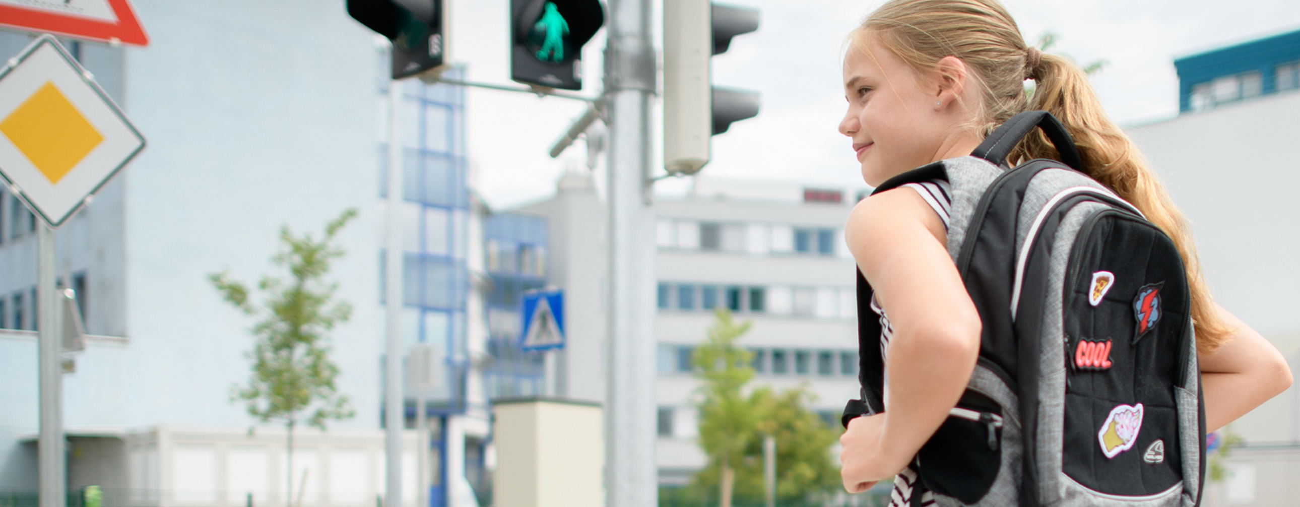 Ein Mädchen mit Schulrucksack steht an einer Fußgängerampel und schaut nach links. Im Hintergrund sind Bürogebäude, Verkehrsschilder und eine Ampel an einer Kreuzung zu erkennen.