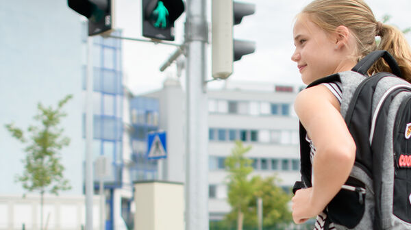 Ein Mädchen mit Schulrucksack steht an einer Fußgängerampel und schaut nach links. Im Hintergrund sind Bürogebäude, Verkehrsschilder und eine Ampel an einer Kreuzung zu erkennen.
