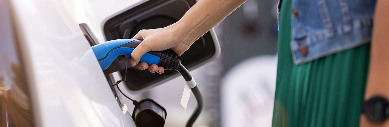 Ein weißes Elektroauto mit Ladekabel, eine Frau hält den Ladestecker in der rechten Hand.