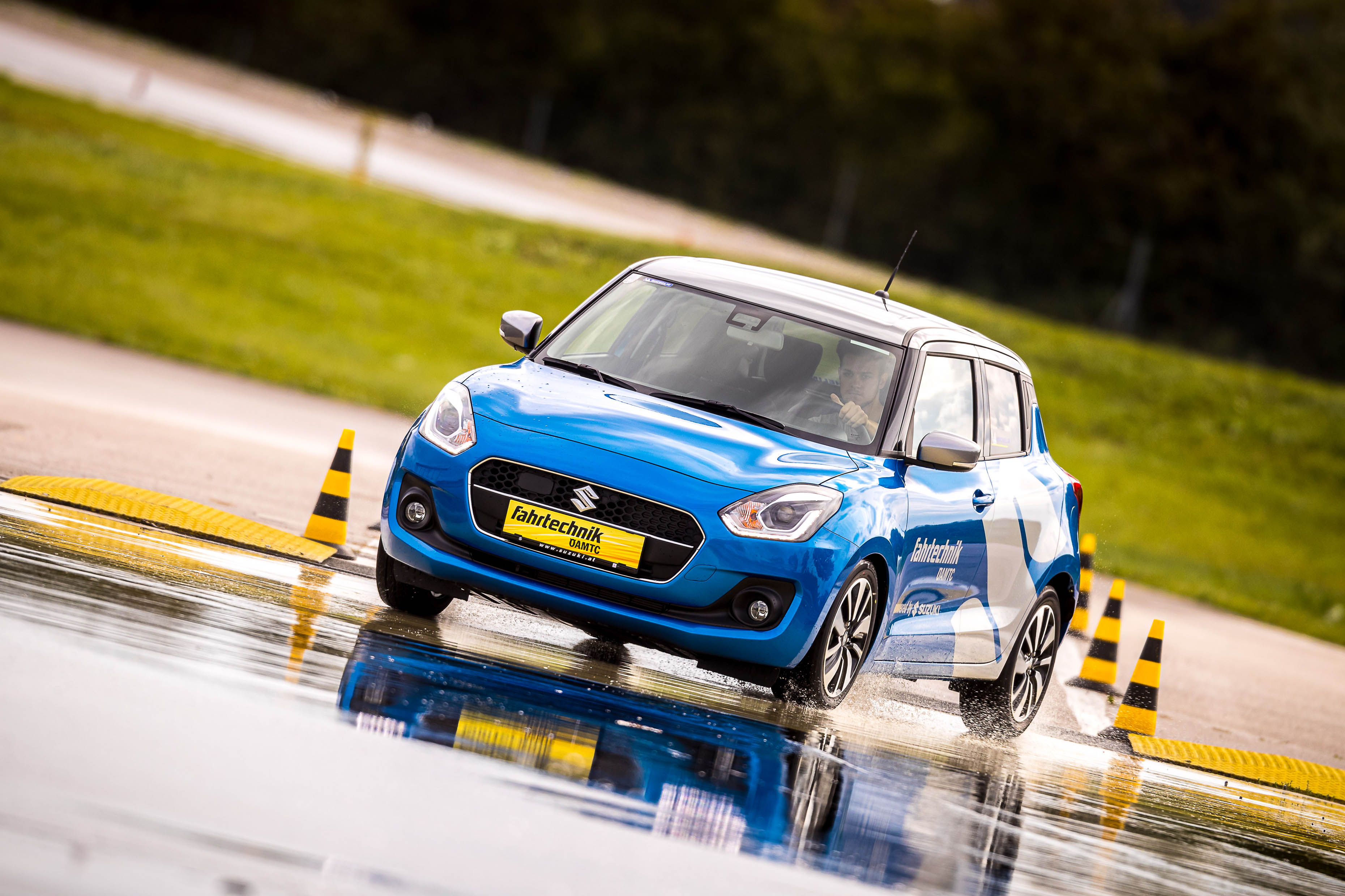 Ein blaues Auto fährt auf nassem Asphalt. Im Hintergrund sind gelb-schwarze Hütchen und grüne Wiesen.