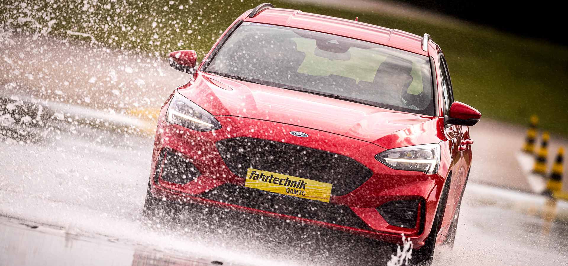 Ein rotes Auto fährt auf nassem Asphalt. Wasser spritzt davon. Im Hintergrund sind gelb-schwarze Hütchen.
