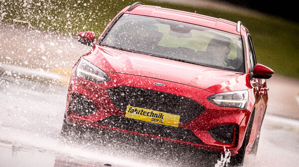 Ein rotes Auto fährt auf nassem Asphalt. Wasser spritzt davon. Im Hintergrund sind gelb-schwarze Hütchen.