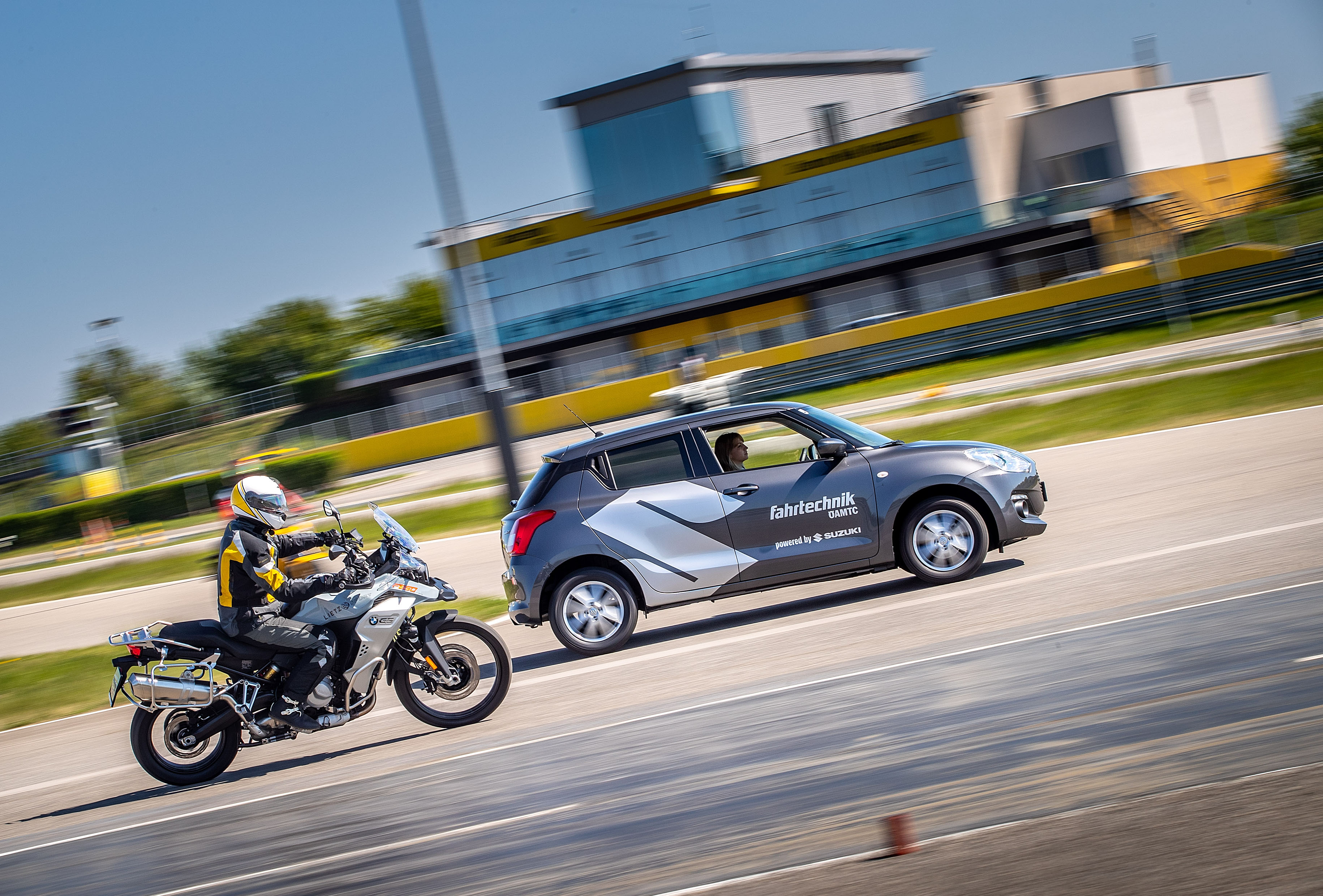 Eine Frau sitzt am Steuer eines grauen Autos. Daneben fährt eine Person auf einem Motorrad mit Motorradhelm und -bekleidung. Im Hintergrund ist ein gelbes Gebäude mit Glasfront.