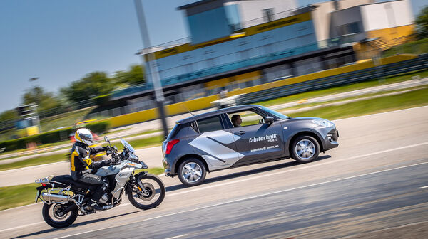 Eine Frau sitzt am Steuer eines grauen Autos. Daneben fährt eine Person auf einem Motorrad mit Motorradhelm und -bekleidung. Im Hintergrund ist ein gelbes Gebäude mit Glasfront.