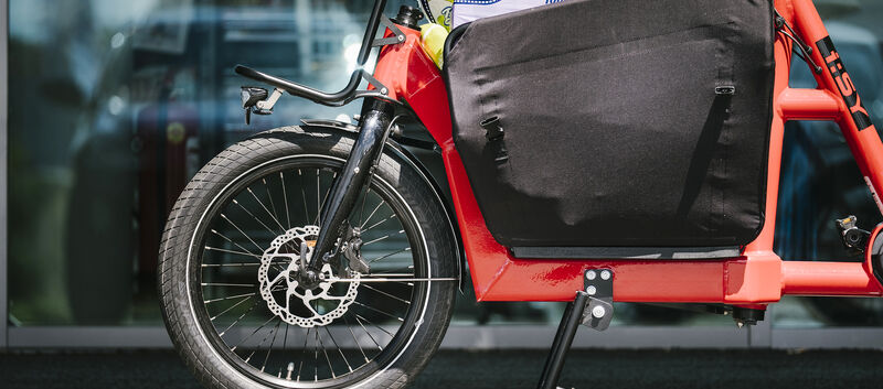 Rotes Lastenfahrrad im Vordergrund, Auto verschwommen im Hintergrund.