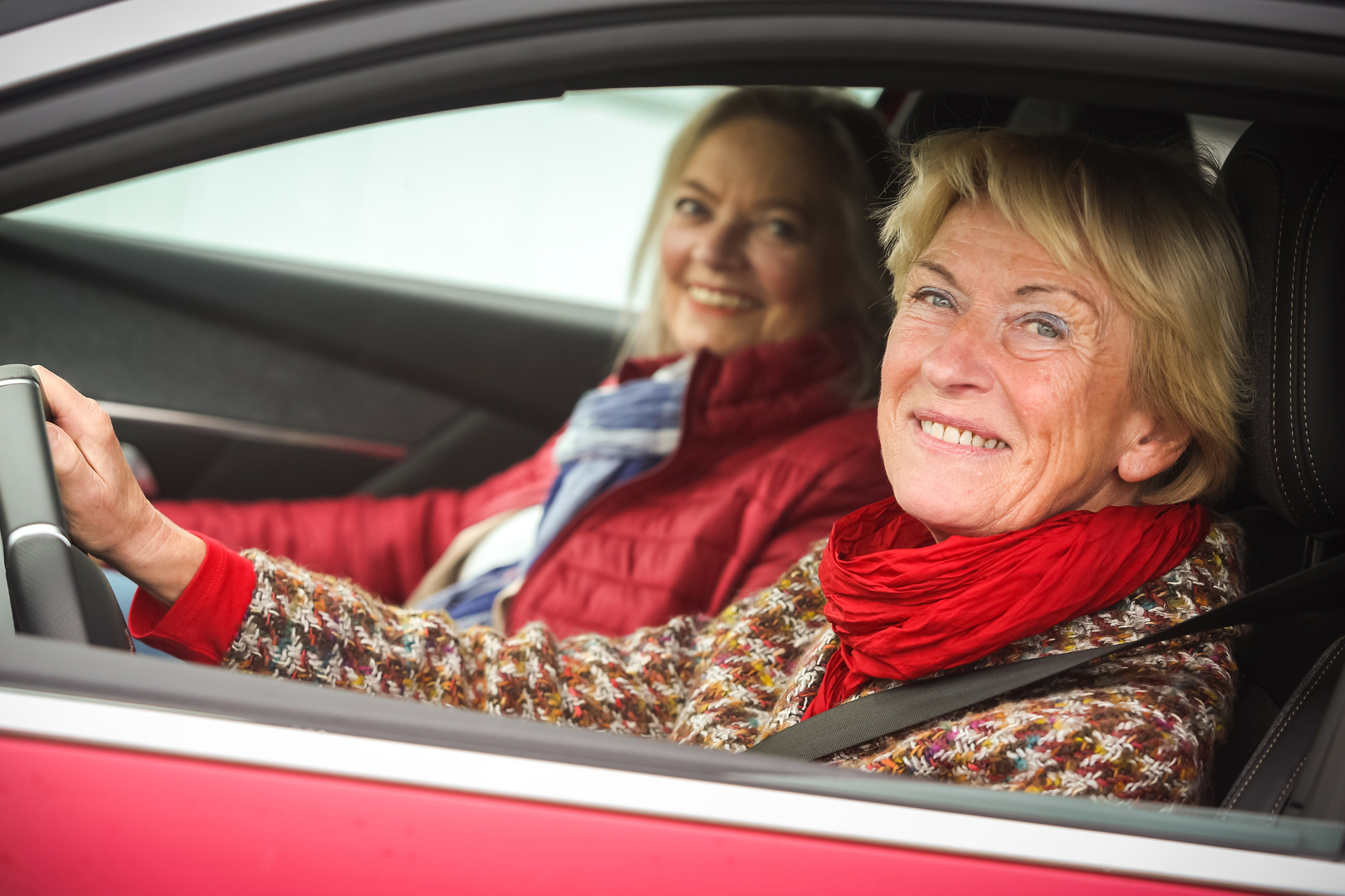 Zwei lächelnde Frauen sitzen in einem roten Auto. Die Fahrerin trägt einen bunten Pullover und einen roten Schal, die Beifahrerin eine rote Jacke und einen blauen Schal.