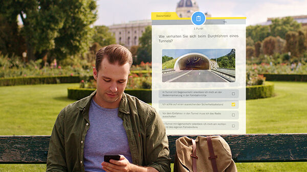 Junger Mann sitzt bei schönem Wetter auf einer Parkbank und blickt konzentriert auf sein Smartphone in der Hand. Im Hintergrund sieht man eine Frage eingeblendet zu einer Tunnel-Durchfahrt der ÖAMTC Führerschein-Test App.