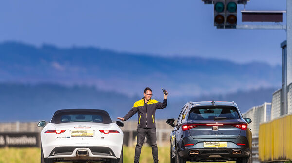 Ein graues Auto und ein weißes Cabrio stehen nebeneinander auf einer Rennstrecke. In der Mitte steht ein Instruktor mit Funkgerät und gibt Anweisungen. Im Vordergrund ist eine Startampel. Im Hintergrund blauer Himmel.