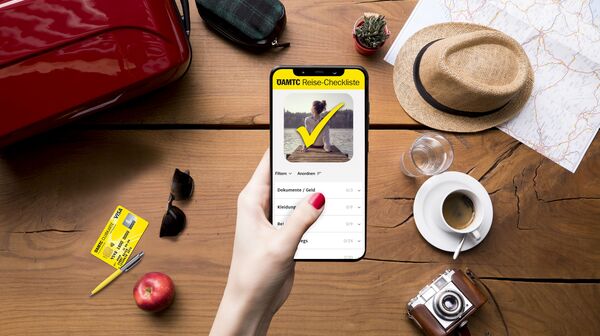 Eine Hand hält ein Smartphone mit der ÖAMTC Reise-Checkliste, umgeben von Reisetasche, Hut, Karte, Kamera, Kaffee und Apfel.