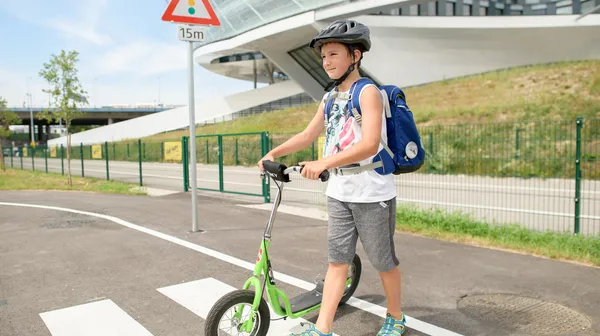 Ein Schulkind mit Schultasche am Rücken und Fahrradhelm am Kopf schiebt einen grünen Scooter über einen Zebrastreifen. Im Hintergrund sind ein asphaltierter Weg und ein großes Bürogebäude.