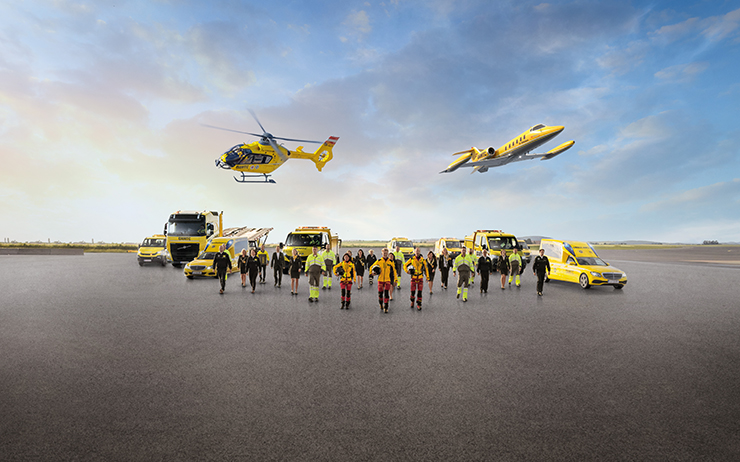 Eine Flotte von gelben Rettungsfahrzeugen und Luftfahrzeugen steht auf einem Flugfeld. Zu sehen ein Krankenwagen, ÖAMTC Pannenfahrzeuge, ein Abschlepp-LKW, ein Rettungshubschrauber in der Luft sowie ein Ambulanzflugzeug. Davor stehen Einsatzkräfte in leuchtender Schutzkleidung. 