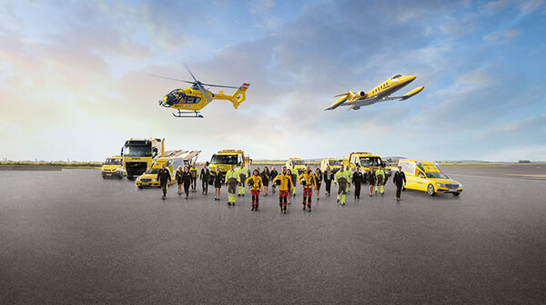 Eine Flotte von gelben Rettungsfahrzeugen und Luftfahrzeugen steht auf einem Flugfeld. Zu sehen ein Krankenwagen, ÖAMTC Pannenfahrzeuge, ein Abschlepp-LKW, ein Rettungshubschrauber in der Luft sowie ein Ambulanzflugzeug. Davor stehen Einsatzkräfte in leuchtender Schutzkleidung.