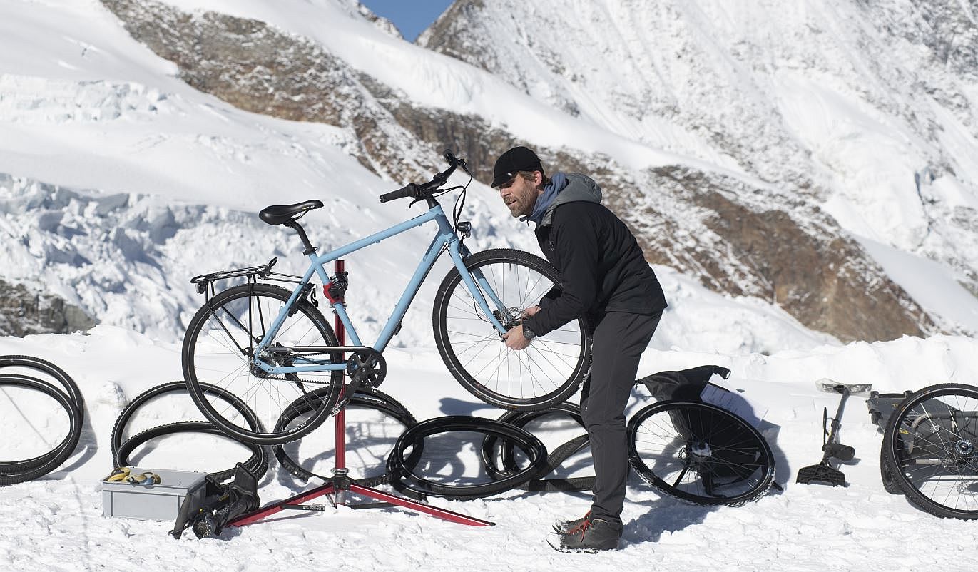 Ein Mann steht im Gebirge im Schnee und repariert ein blaues Fahrrad auf einem Fahrradständer. Er ist von mehreren Fahrradreifen umgeben und im Hintergrund sind schneebedeckte Berge zu sehen.