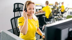 Auf dem Bild sieht man eine Frau in einem kurzärmeligen gelben T-Shirt. Am Ärmels steht ÖAMTC. Sie sitzt vor einem Bildschirm und hat ein Headset auf.