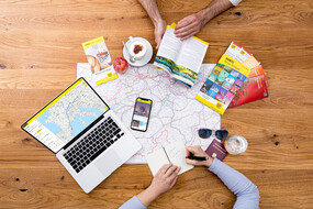 Zwei Personen planen eine Reise, nutzen Karten, Reiseführer, einen Laptop und ein Smartphone.
