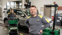 Ein männlicher Kfz-Techniker in Dienstkleidung (grau-gelber Pullover) steht mit seiner Kollegin (ebenfalls Kfz-Technikerin) in der Werkstatt. Er lacht in die Kamera, die weibliche Person arbeitet konzentriert. Im Hintergrund ist ein grauer PKW zu sehen.
