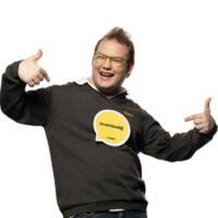 Ein junger Kollege mit Brille in Schalterdienstkleidung (grauer Pullover, weißes Hemd darunter) lacht in die Kamera und zeigt mit beiden Händen auf sich selbst.